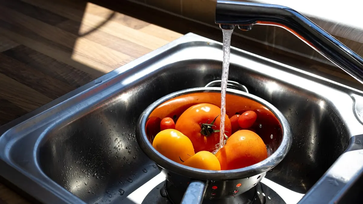 Pia de inox com água correndo da torneira sobre bacia de tomates